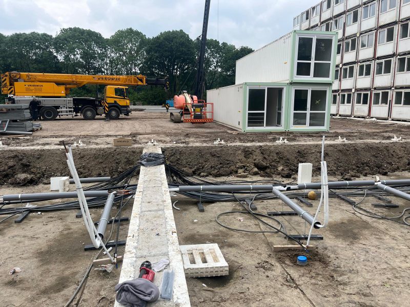 Plaatsing van witte containerwoningen met een gele kraan, terwijl kabels en leidingen op de voorgrond worden aangelegd